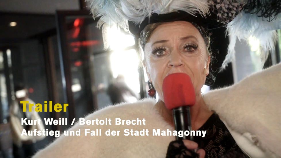 TITLE: Kurt Weill / Bertolt Brecht: Aufstieg und Fall der Stadt Mahagonny
URHEBER: Ruth Tromboukis Dieses Foto ist das Standbild zum Stücktrailer: Leokadja Begbick begrüßt das Publikum in der Netzestadt Mahaonny. Ein rotes Handmikrofon verstärkt ihre Stimme, so dass alle im Foyer ihren Ruf "Willkommen in Mahagonny, willkommen zu Hause" hören können.