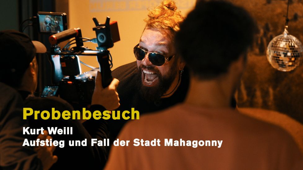 TITLE: Ein Probenbesuch bei Aufstieg und Fall der Stadt Mahagonny
URHEBER: Ruth Tromboukis Auf diesem Standbild zum Probenbesuch-Teaser ist Robert Gleadow, der Darsteller des Dreieinigkeitsmoses. Er lacht diabolisch in die Kamera. Hinter ihm eine kleine Disko-Kugel. Auf dem Standbild stehen die Worte: Probenbesuch, Kurt Weill, Aufstieg und Fall der Stadt Mahagonny.