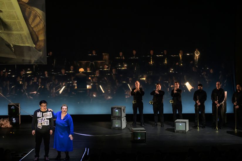 TITLE: Aida
URHEBER: Bettina Stöß Radames träumt sich in die Rolle eines erfolgreichen Heerführers. Amneris heftet ihm auf den Pullover Zeitungsbilder von Kriegsverbrechen. Rechts stehen die Musiker des Orchesters mit Königstrompeten für den Triumphmarsch.