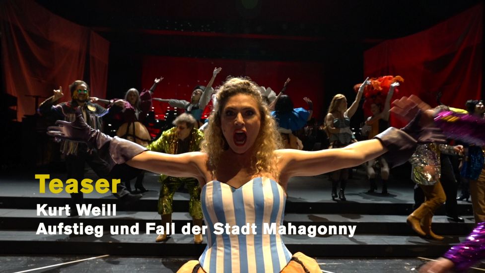 TITLE: Aufstieg und Fall der Stadt Mahagonny (Ein Teaser)
URHEBER: Ruth Tromboukis Dieses Bild ist das Standbild zum Teaser. Jenny Hill in einer blau-weißen Korsage breitet die Arme weit zu beiden Seiten aus. Hinter ihr weitere Darsteller und das Orchester der Deutschen Oper Berlin.