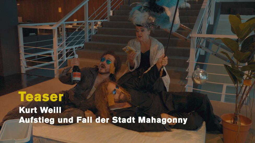 TITLE: Aufstieg und Fall der Stadt Mahagonny (Ein Teaser)
URHEBER: Ruth Tromboukis Am Fuße einer Treppe im Foyer lagern Witwe Begbick, Fatty und Dreieinigkeitsmoses mit Bündeln Gelds, mit Mahagonny-Sekt, einer Topfpflanze und einer Diskokugel am Besenstiel. Es ist das Standbild zu einem kurzen Teaser.