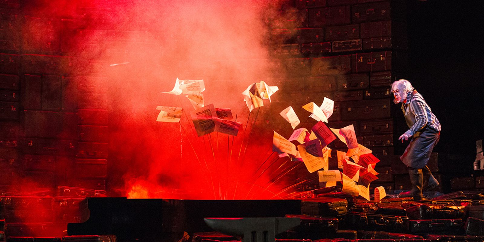 Siegfried - ©2021 ©Bernd Uhlig Mime steht auf einer Anhäufung von Koffern. Er blickt auf den Flügel, aus dessen Inneren zahllose Notenblätter aufsteigen. Die Bühne ist in leuchtend roten Neben getaucht.