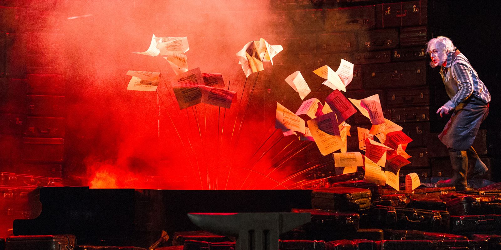 Der Ring des Nibelungen - ©Bernd Uhlig Auf dem Foto eine Szenenimpression aus SIEGFRIED. Mime steht rechts im Bild in einer Schmiedeschürze und mit schweren Stiefeln. Aus dem Flügel, der als ein Requisit alle Teile des RINGs durchzieht, steigen wehend Notenblätter auf. Die Szenerie wird bestimmt durch dichten roten Nebel. Die Bühnenbegrenzung nach hinten bilden die Stapel an Koffern, die ebenfalls als durchziehendes Bühnenbildteil alle vier Werke des RINGs prägen.
