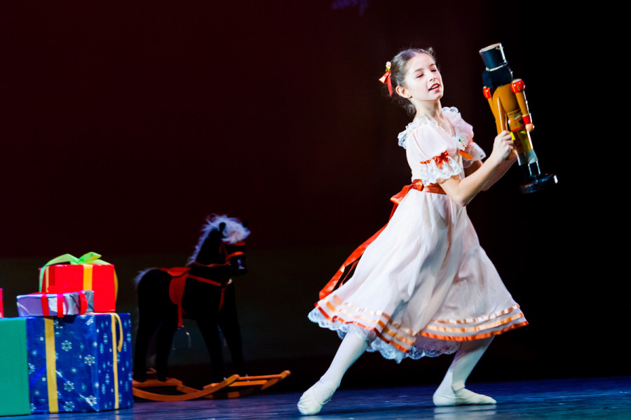 Kinder tanzen: Der Nussknacker - © Yan Revazov / KBKB Verpackte Weihnachtsgeschenke und ein Schaukelpferd bilden die Kulisse. Glücklich hält Klara den Nussknacker in den Händen.