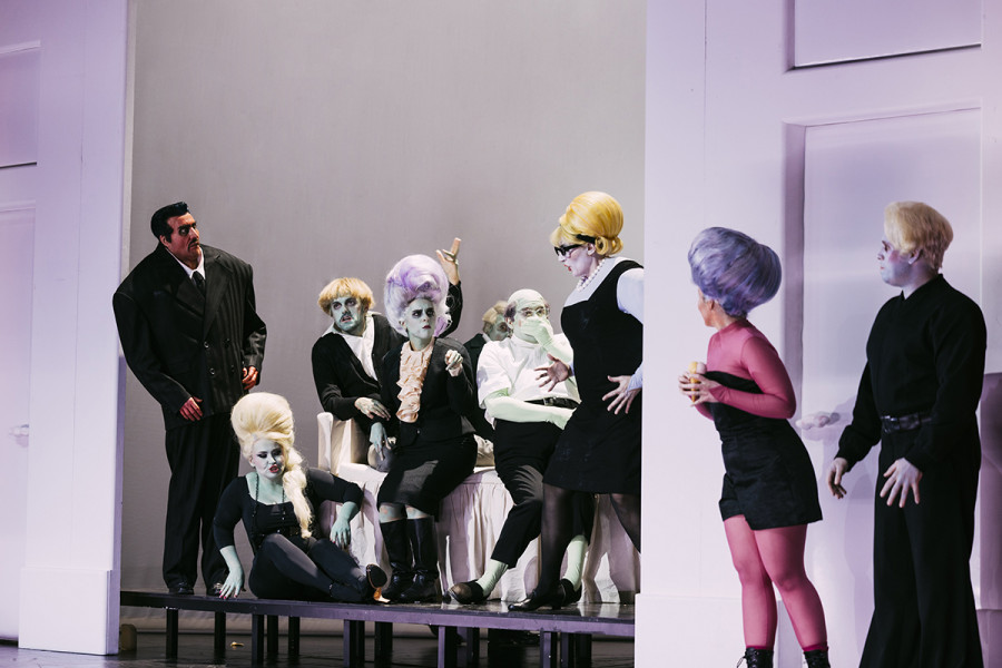 Gianni Schicchi - © Eike Walkenhorst Die Darsteller*innen haben grün oder gelb geschminkte Gesichter und Hände. Die Kostüme sind schwarz/weiß. Die Verwandtschaft - Marco, Zita, Simone, Nella, La Ciesca - umstehen das Bett des Buoso Donati. Links neben dem Kabinett mit Buosos Sterbebett das Liebespaar Lauretta, Rinuccio.