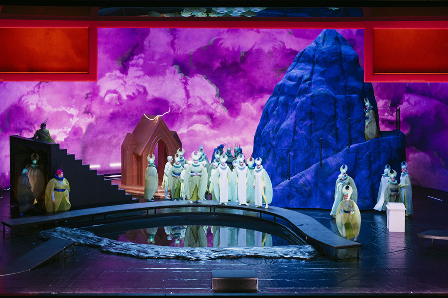 Bühnentotale mit allen Bestandteilen: Ein blauer Felsen, die orangene Kapelle, eine Treppe und das Wasserbecken. Verschiedene Gruppen von Nonnen verteilen sich in diesem Bild.