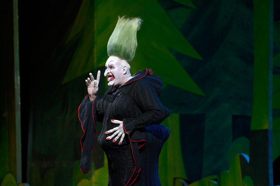 Hänsel und Gretel - © 2014 // Bettina Stöß Die Hexe (sie wird von einem Tenor gespielt und gesungen) steht zaubernd auf ihrer Märchen-Lichtung. Sie trägt ein nachtschwarzes Plisseekleid mit leuchtend roten Saumnähten. Ihre Haare sind leuchtend grün wie ihr Märchenwald und stehen steil nach oben. Strukturen im Kostüm bilden unnatürlich große Brüste, einen spitzen Bauch und einen riesigen runden Po. Ihre Lippen und Fingernägel sind knallrot.