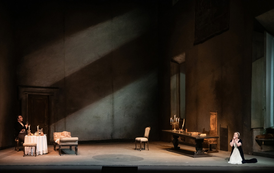 Tosca - © 2023, Bettina Stöß Scarpia sitzt auf einem Polsterstuhl am Tischchen, Tosca kniet flehend in der anderen Raumälfte. Sie singt ihre berühmte Arie "Vissi d’arte".