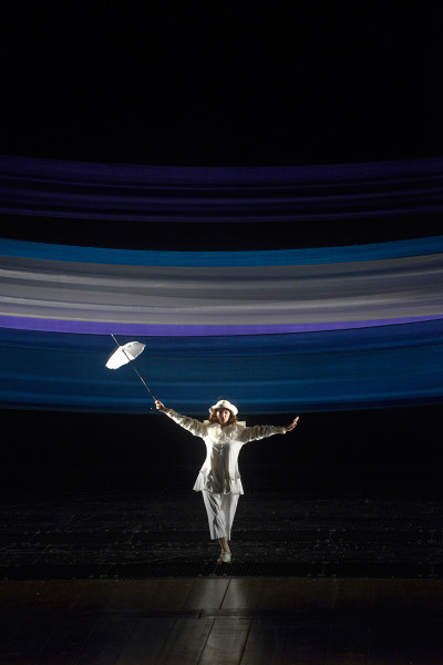 Die Zauberflöte - © Bettina Stöß Ein blau-lila-weißes Tuch spannt sich über den Horizont. Aus dem Bühnenhintergrund tänzelt Papagena nach vorne. Sie trägt ihr weißes Kolombine-Kostüm mit weißem Hütchen und weißem Schirm. Sie sieht nun jung aus.