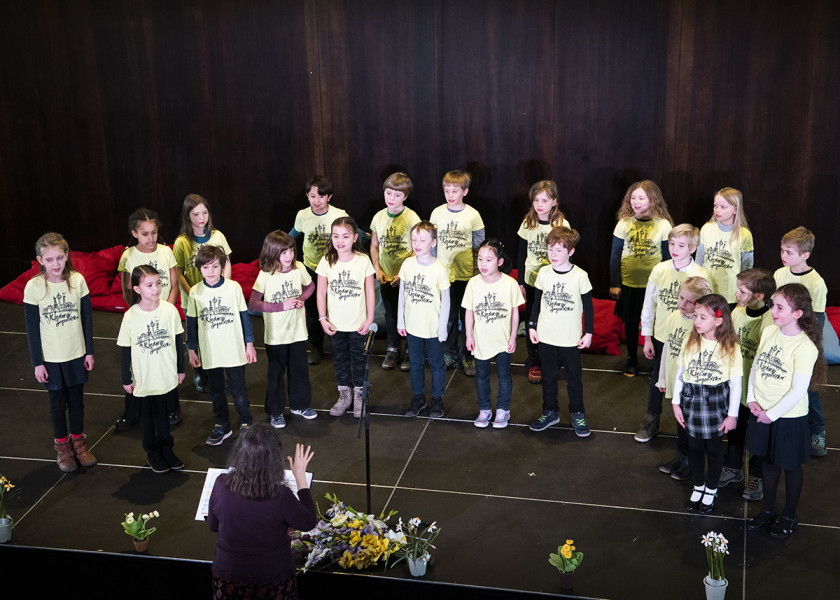 Frühlingssingen - © S.I. In einem Halbkreis um seine Dirigentin Rosemarie Arzt stehen die Kinder des Kleinen Chores. Am Bühnenrand stehen Frühlingsblumen.