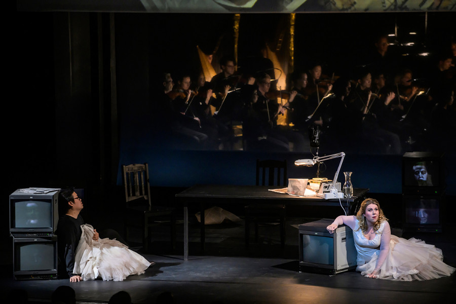 Aida - © Bettina Stöß Radames lehnt sich an zwei Bildschirme auf der Bühne, Aida kniet in einem Brautkleid am Boden. Hinter ihnen Radames' Schreibtisch, dahinter das Orchester der Deutschen Oper Berlin.