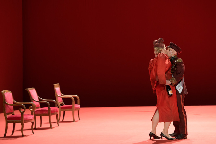 A Midsummer Night’s Dream - © Bettina Stöß Die Bühne ist in rot getaucht. Drei rote Armsessel stehen diagonal. Das Herzogspaar Theseus und Hippolyta küssen sich. Ihre Kostüme sind ebenfalls rot. Sie trägt ein Bleistiftkleid, das an die Moder der 30er Jahre erinnert, er eine Uniform.