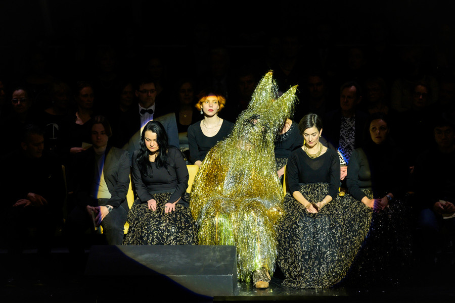 Rigoletto - © Bettina Stöß Rigoletto sitzt im Goldlametta-Hasenkostüm in den Stuhlreihen des Eröffnungsbühnenbildes. Der Saal der Deutschen Oper Berlin mit seinen typischen gelben Sitzbezügen ist mit Parkett und erstem Rang gespiegelt. In diesem zweiten Saal sitzen Chor und Statisterie.