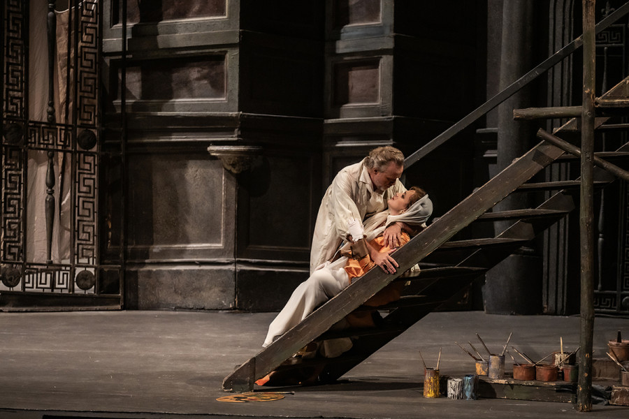 Tosca - © 2023, Bettina Stöß Auf der Holztreppe, die zum Fresko führt, an dem Cavaradossi arbeitet, liegen eng umschlungen Tosca in den Armen Cavaradossis. Unter dem Treppenpodest stehen viele Farbtöpfe.