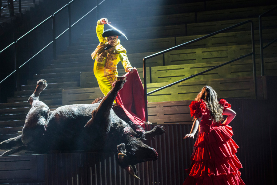Im Bildvordergrund der getötete Stier. Über ihm in stolzer Haltung Escamillo, sein Langmesser gezückt. Daneben Frasquita in einem ebenso ausladend roten Volantkleid wie Carmen.
