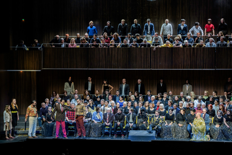 Monterone verflucht Rigoletto: Monterone in einem gepflegten Herrenoutfit, rechts kniend neben ihm Rigoletto im goldenen Lametta-Narren-Gewand, die Kappe mit den Hasenohren hat er inzwischen abgenommen. Der Hofstaat lauscht der Verfluchung interessiert.