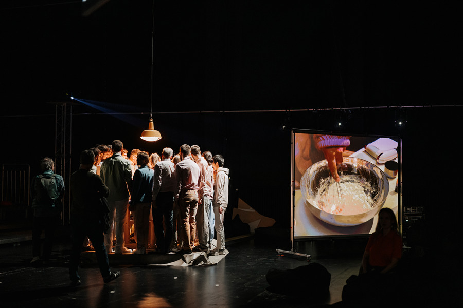 Satisfactionaction - © Nancy Jesse Die Chormitglieder wenden sich von einem Videoscreen ab. Auf dem Screen sieht man einen Mixer, der Zutaten in der Schüssel vermengt.