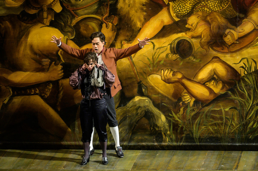 Le nozze di Figaro - © Bettina Stöß Der Page wird delegiert zur Armee des Grafen, denn er soll aus dem Wege. Figaro schildert Cherubino amüsiert sein künftiges Aufgabengebiet: Kampffeld statt Hofleben. Hinter den beiden, die nah an der Rampe stehen, ist ein Vorhang mit einem Schlachtengemälde.