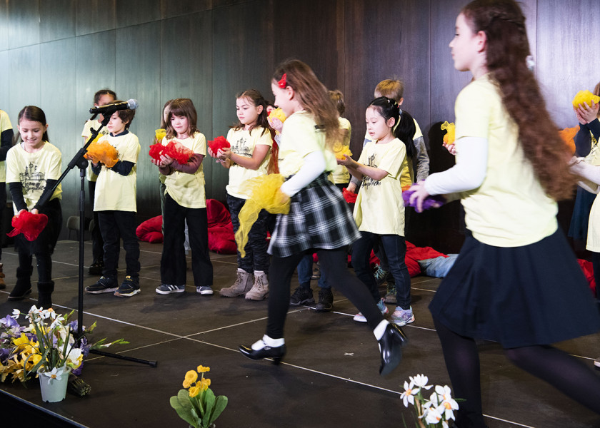 Frühlingssingen - © S.I. Der Kleine Chor des Kinderchores bei seinem Frühlingssingen: Die Kinder singen und tanzen auf der Foyerbühne in hellgelben T-Shirts. Verstreut sind einige Narzissen und Frühblüher.