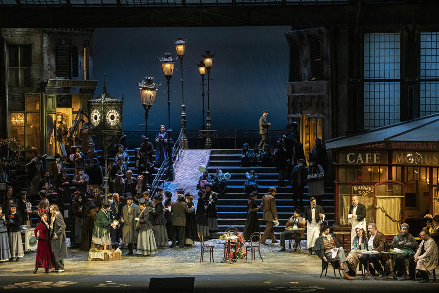 La Bohème - © Bettina Stöß Der Platz hat sich in der Mitte ein wenig gelehrt. Links am Portal stehen Marcellos ehemalige Geliebte Musetta mit ihrem neuen Verehrer, dem reichen Alcindoro. Rechts, vor dem Café Momus sitzen Mimì, Rodolfo, Marcello, Colline und Schaunard.