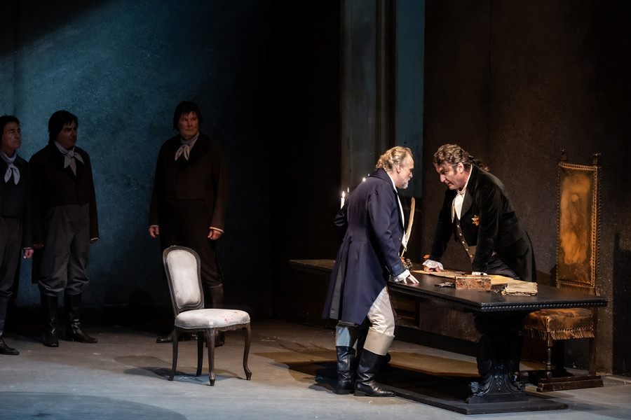 Tosca - © 2023, Bettina Stöß Am Schreibtisch stehen sich Cavaradossi und Scarpia gegenüber. Drohend beugen sie sich gegeneinander. Im Hintergrund warten die Schergen des Scarpia auf Befehle ihres Herrn.