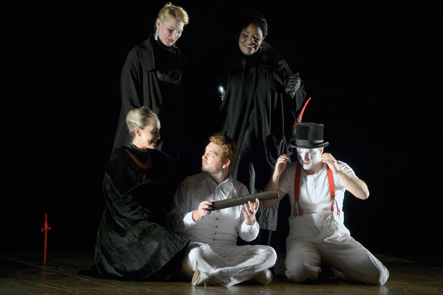 Die Zauberflöte - © Bettina Stöß Auf dem Foto umstehen die drei Damen der Königin der Nacht Tamino und Papageno. Gerade haben sie Tamino die magische Flöte überreicht. Die Damen tragen schwarze sehr elegante Gewänder, sind opulent geschmückt und haben Abendfrisuren. Tamino trägt weißes Hemd, Weste, Hose und Schuhe. Sein Gesicht ist hautfarben. Papageno trägt weiße Hose und T-Shirt sowie seinen schwarzen Zylinder mit roter Feder. Sein Gesicht ist weiß geschminkt.