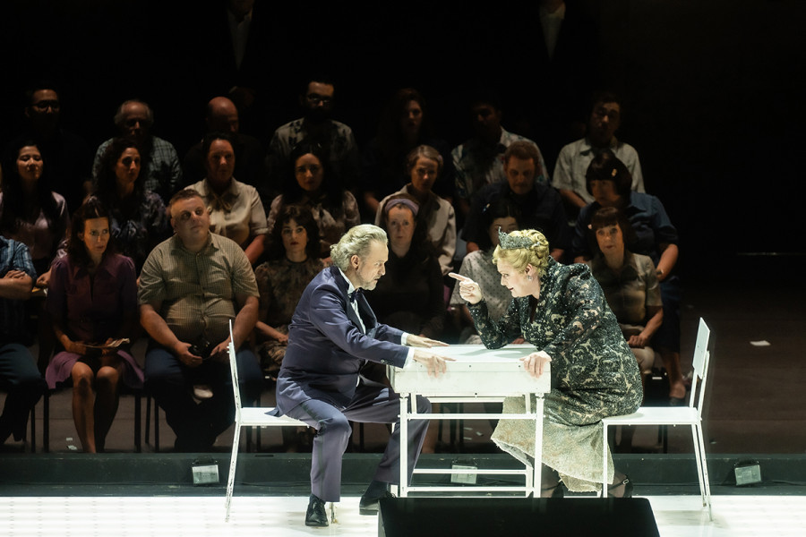 Turandot - © 2023 // Bettina Stöß Calaf sitzt an dem kleinen weißen Tisch Turandot gegenüber. Mit beiden Händen hält er sich am Tisch fest. Turandot zeigt herausfordernd mit dem Zeigefinger der rechten Hand auf ihn. Im Hintergrund das Volk, das mit Spannung der Rätselbefragung folgt.