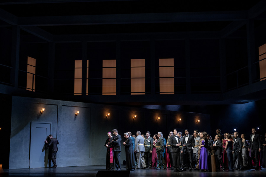 Simon Boccanegra - © Bettina Stöß Die Bühne ist horizontal und vertikal zweigeteilt. Eine mobile Wand trennt den unteren Bereich der Bühne, eine Fenster-Galerie verläuft oberhalb der Raummitte. Die Wand steht diagonal auf der Bühne. Im rechten Bereich der Bühne steht der Chor in Galauniformen und Abendgarderobe. Ein Mann lauscht an einer Tür, die ins Gemach hinter der Wand führt.