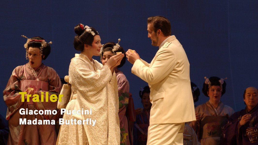 Giacomo Puccini: Madama Butterfly - © Ruth Tromboukis Dieses Foto ist das Standbild zu Videotrailer mit Szeneneindrücken aus Pier Luigi Samaritanis Inszenierung zu MADAMA BUTTERFLY aus dem Jahre 1987. Cio-Cio-San und Pinkerton stehen sich verliebt gegenüber. Sie in weißem Kimono mit Blüten im Haar, er in weißer Galauniform.