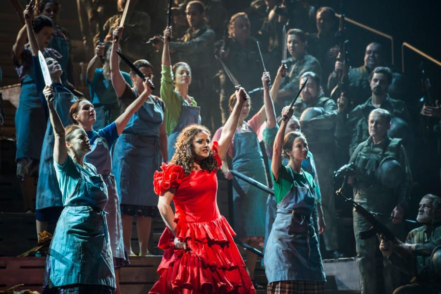 Carmen - © Marcus Lieberenz Carmen im roten Kleid, der Chor in Arbeitskleidung. Die Frauen halten Messer nach oben, die Männer bedrohen sie mit Maschinengewehren.