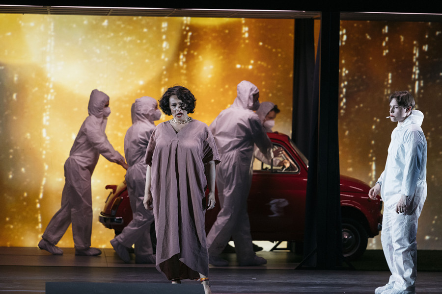 Ángeles Blancas Gulin als Lucia mit Eric Naumann als Wissenschaftler im Vordergrund des Bildes, im Hintergrund schieben vier maskierte Wissenschaftler den roten Fiat von der Bühne. Goldenes Feuerwerk bildet den Horizont.