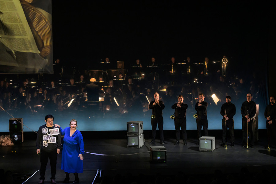 Aida - © Bettina Stöß Radames träumt sich in die Rolle eines erfolgreichen Heerführers. Amneris heftet ihm auf den Pullover Zeitungsbilder von Kriegsverbrechen. Rechts stehen die Musiker des Orchesters mit Königstrompeten für den Triumphmarsch.