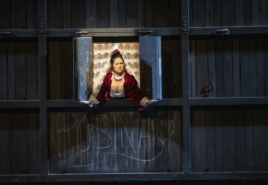 Il barbiere di Siviglia - © Bettina Stöß Berta, die Gesellschafterin Rosinas, schaut wütend aus dem Fenster. Sie sieht nicht, dass Almaviva mit Kreide ein großes Herz an den Wohnwagen gemalt und in Großbuchstaben Rosina geschrieben hat.