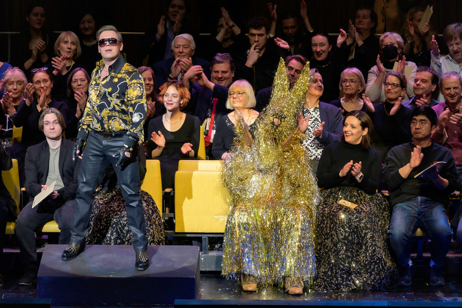 Rigoletto - © Bettina Stöß Der Herzog steht auf dem Souffleurkasten und singt ins Publikum. Hinter ihm im gespiegelten Saal sitzt der Hofstaat, in der Mitte Rigoletto im Lametta-Kostüm.