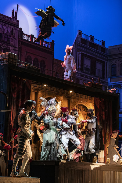 Es ist das große Finale der Oper: Die Protagonist*innen tanzen ausgelassen. Im Vordergrund Figaro in einem typischen Harlekin-Kostüm der Commedia dell'arte.