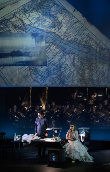 Aida - © Bettina Stöß Aida sitzt bei Radames am Schreibtisch. Auf der Videoleinwand ist eine Landkarte des Nilverlaufes und eine Postkarte mit eine Nillandschaft im Sonnenuntergang.