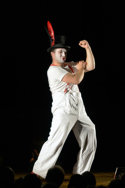Die Zauberflöte - © Bettina Stöß Auf dem Bild ist Papageno in herausfordernder Pose. Er trägt eine weiße Hose mit roten Hosenträgern und ein weißes T-Shirt. Er trägt einen schwarzen Zylinder mit einer roten Feder. Sein Gesicht ist weiß geschminkt.