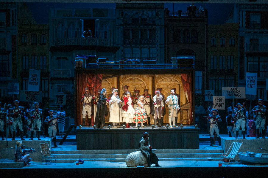 Der Barbier von Sevilla - © 2013 // Marcus Lieberenz Das große Finale des 1. Aufzugs. Die Straße mit dem Theaterwagen ist in blaues Mondlicht getaucht. hell beleuchtet ist das Innere des Theaterwagens. Basilio, Bartolo, Berta, Rosina, Almaviva in Verkleidung als Soldat, Figaro und einige weitere Soldaten bevölkern den Wagen. Links und rechts neben dem Wagen demonstrieren Stadtbewohner mit Schildern und Parolen wie: "Make Holiday not war", "Keine Gewalt", "Freiheit für die Kunst" ...