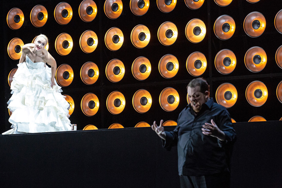 Tristan und Isolde -   © Bernd Uhlig  Tristan und Isolde sind sich in Liebe verfallen. Isolde steht im weißen Braukleid auf dem Podest, Tristan in schwarzer Hemd-Hose-Kombination steht vor ihr am Boden.