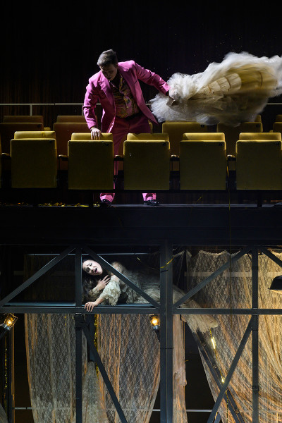 Rigoletto - © Bettina Stöß Die Rückseite der nach hinten offenen Unterbühnen-Konstruktion ist durch altmodische Gardienen (Stores) verhängt. Gilda trägt ein Kleid aus ebendiesem Gardinenstoff. Der Herzog steht in den nun aufgefahrenen Sitzreihen, auffällig in Pink gekleidet und wieder mit gemustertem Seidenhemd und lockt Gilda mit einem Brautkleid.