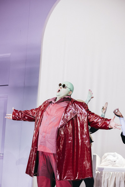 Gianni Schicchi - © Eike Walkenhorst Gianni Schicchi mit rotem Mantel und rotem Anzug. Der Kopf ist grün. Er trägt Sonnenbrille.