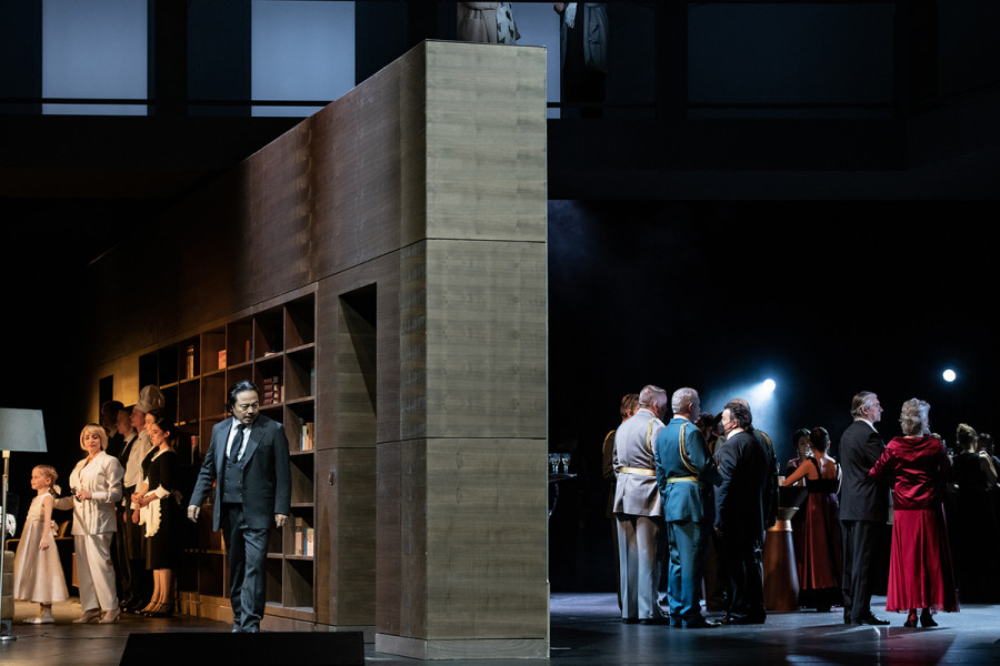 Simon Boccanegra - © Bettina Stöß Eine schwere graue Wand teilt die Bühne. Während auf der linken Seite ein Privatraum erkennbar ist - Dienstpersonal und die Familie des Jacopo Fiesco sind das dramatische Personal -, ist auf der rechten Seite ein öffentlicher parlamentarischer Raum. Dort stehen Besucher in offizieller Kleidung wie Uniformen und Abendrobe.