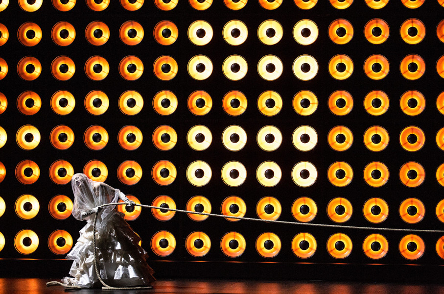 Tristan und Isolde -   © Bernd Uhlig  Vor einer Wand aus zahllosen Lampen zieht Isolde an einem Seil. Sie trägt ein Brautkleid.