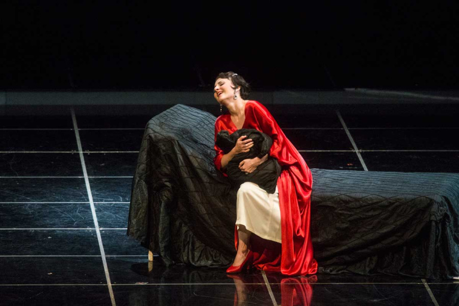 La Traviata - © Marcus Lieberenz Sehnsüchtig sitzt Violetta auf einem Bett, das mit einer schwarzen Seidendecke abgedeckt ist. Über einem weißen Nachthemd trägt sie einen roten Seidenmantel und rote Riemchenpumps. Sie presst sich ein Kissen gegen die Brust.