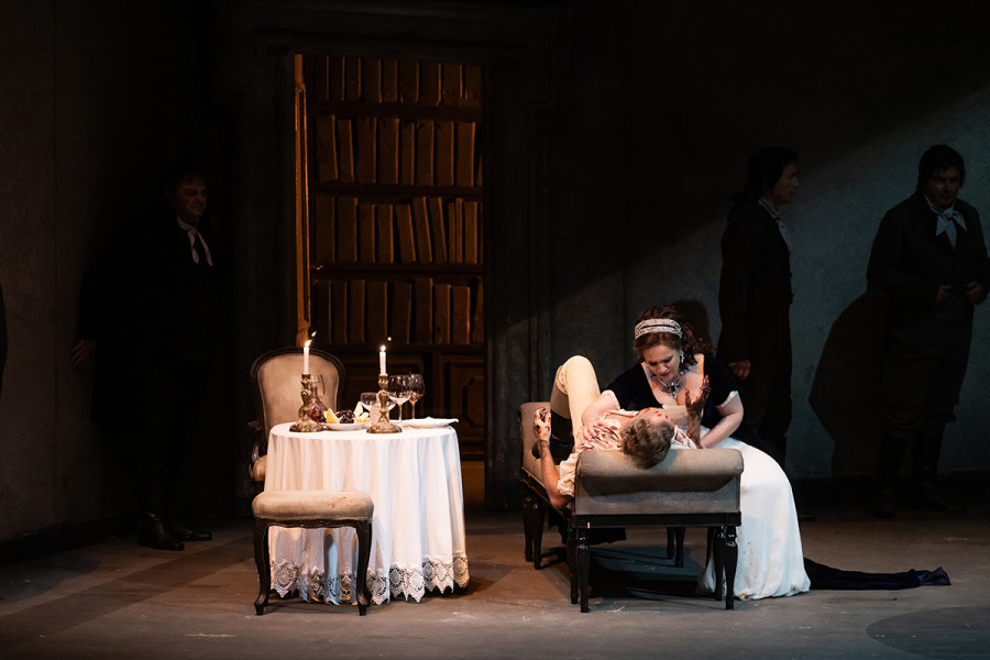 Tosca - © 2023, Bettina Stöß Von der Folter gezeichnet liegt Cavaradossi auf der Ottomane, Tosca beugt sich sorgend und tröstend über ihn. In den dunklen Ecken des Raumes beobachten dies die Schergen des Scarpia.