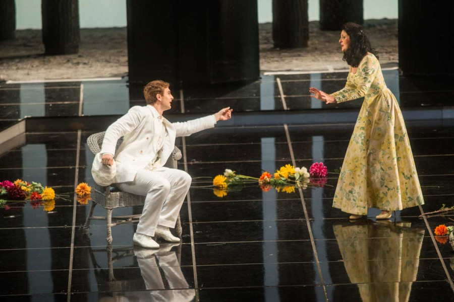 La Traviata - © Marcus Lieberenz Alfredo und Violetta leben auf dem Land. Er sitzt in einem leichten Korbstuhl. Er trägt einen weißen Leinenanzug, sie ein mit Blumen bedrucktes Kleid. Auf dem Boden liegen verstreut Blumen.