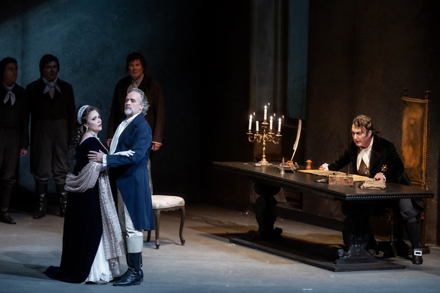 Tosca - © 2023, Bettina Stöß Mit stolzem Blick steht Cavaradossi bei Tosca, die verängstigt wirkt. Scarpia am Schreibtisch sitzend hat den Blick böse auf das Paar gerichtet.