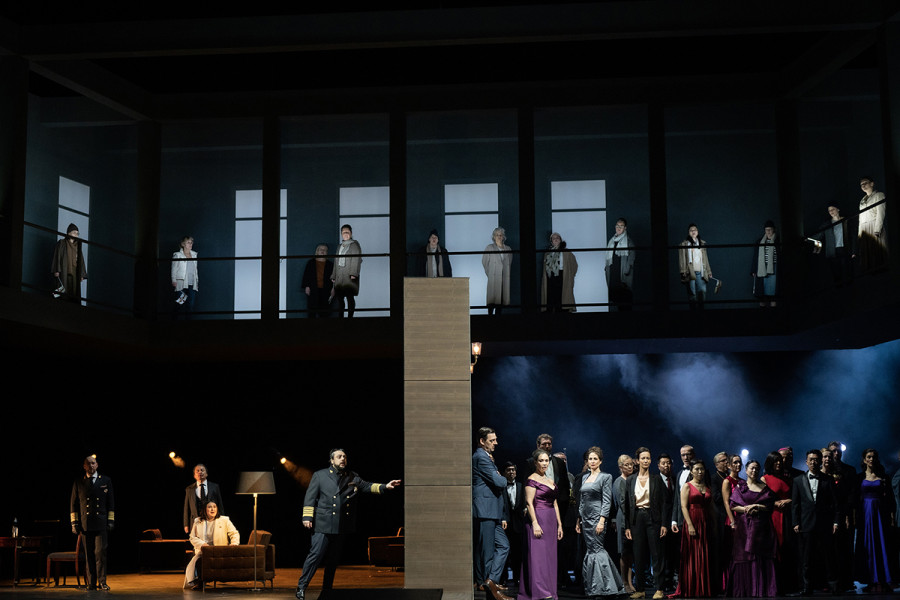 Simon Boccanegra - © Bettina Stöß Eine schwere graue Wand teilt die Bühne. Während auf der linken Seite ein Privatraum erkennbar ist - Dienstpersonal und die Familie des Jacopo Fiesco sind das dramatische Personal -, ist auf der rechten Seite ein öffentlicher parlamentarischer Raum. Dort stehen Besucher in offizieller Kleidung wie Uniformen und Abendrobe.