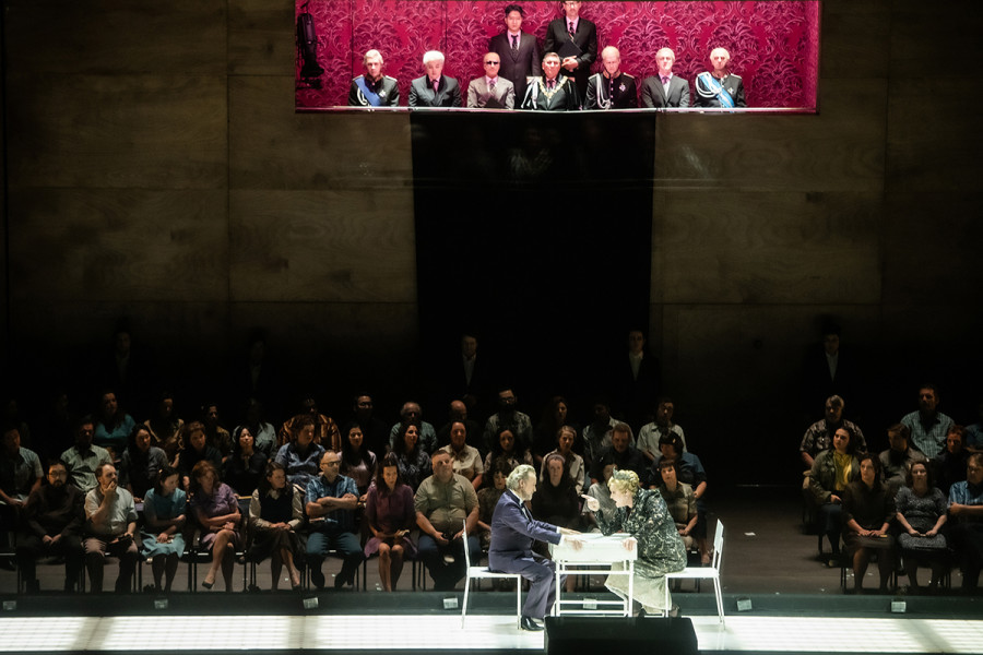 Turandot - © 2023 // Bettina Stöß Calaf sitzt an dem kleinen weißen Tisch Turandot gegenüber. Mit beiden Händen hält er sich am Tisch fest. Turandot zeigt herausfordernd mit dem Zeigefinger der rechten Hand auf ihn. Im Hintergrund das Volk, das mit Spannung der Rätselbefragung folgt. Oben in der Wand ist die Tribüne für die Regierungsriege wieder mit Würdenträgern gefüllt. Auch sie verfolgen die Befragung.