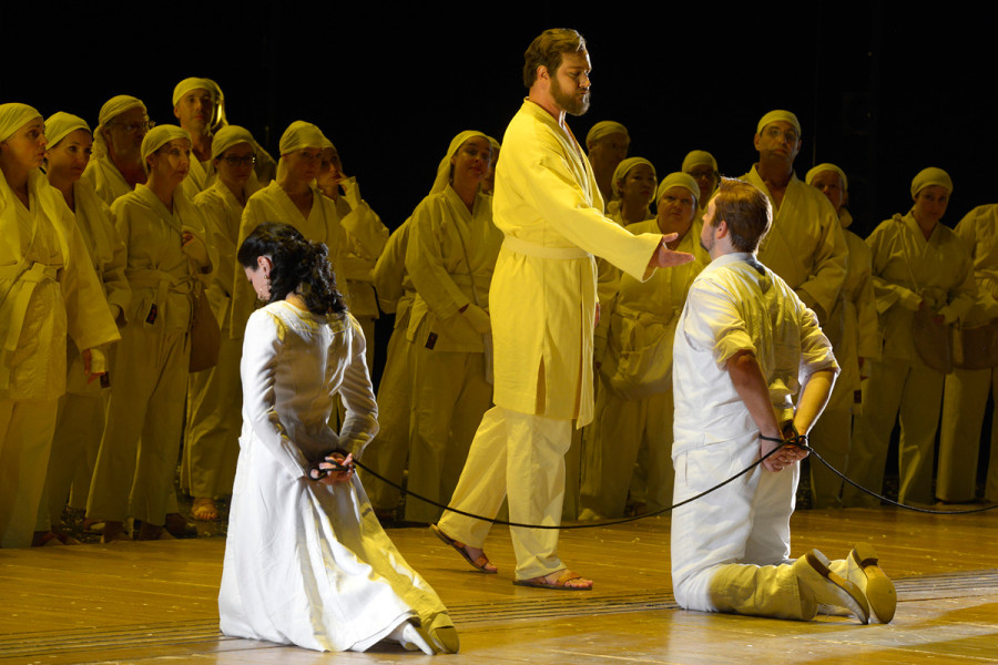 Die Zauberflöte - © Bettina Stöß Pamina und Tamino sind an den Händen gefesselt und knien mit dem Rücken zum Publikum. Vor ihnen Sarastro, dahinter der Chor: Pamina und Tamino gekleidet in weiß, Sarastro und der Chor in sonnengelb.
